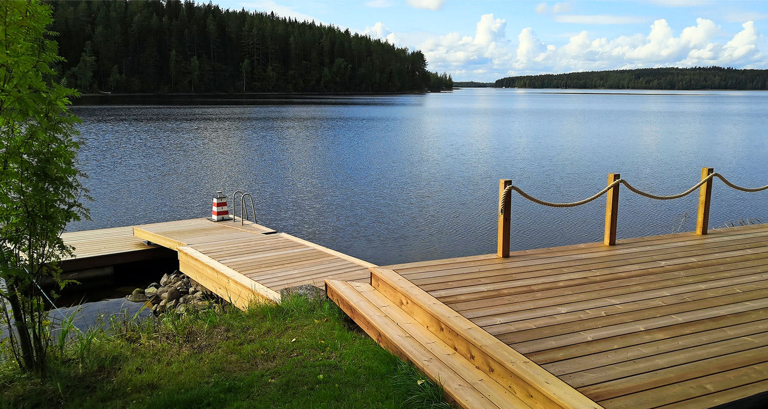Puulaituri sinisen järven rannassa, köysikaiteet ja vihreä metsäranta taustalla