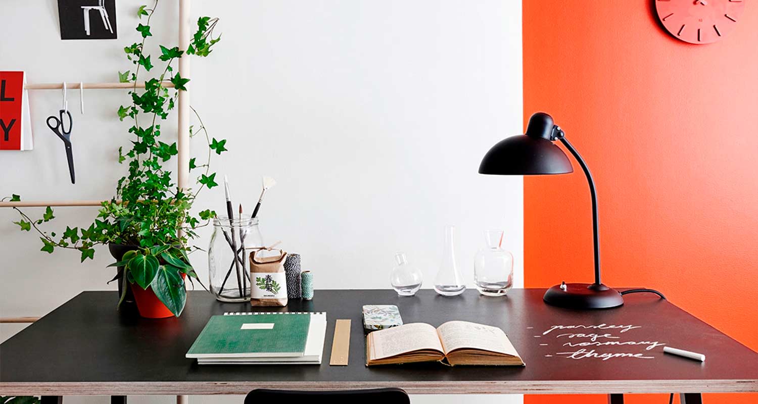 Musta työpöytä valko-oranssiseinän edessä: viherkasvi, siveltimiä, avoin vihko ja musta lamppu