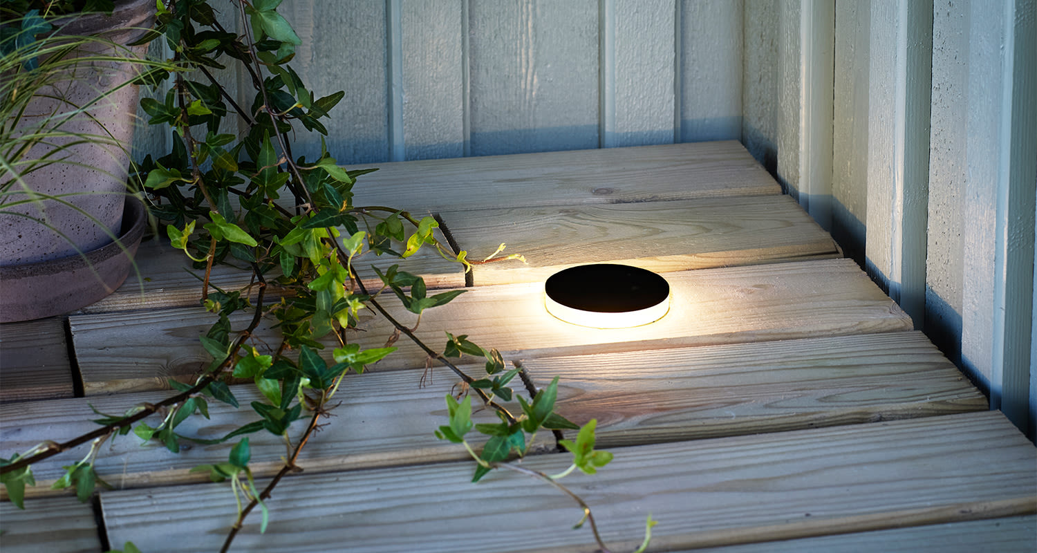 Pyöreä musta LED-valaisin lämpimällä valolla puuterrassilla, vierellä vihreä köynnös.