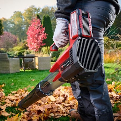 Henkilö puhaltaa punamustalla akkulehtipuhaltimella syksyn lehtiä nurmikolta puutarhakalusteiden edessä.