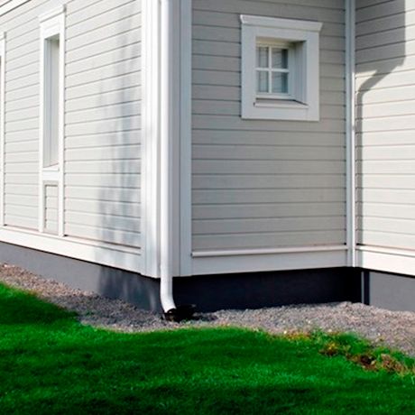 Vaaleaharmaan puutalon kulma, valkoinen ränni, pieni ikkuna ja vihreä nurmikko.