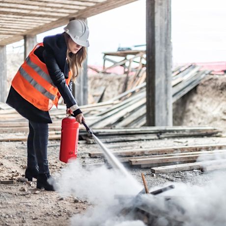 Rakennustyömaalla oranssiliivinen henkilö kypärä päässä sammuttaa savua punaisella sammuttimella