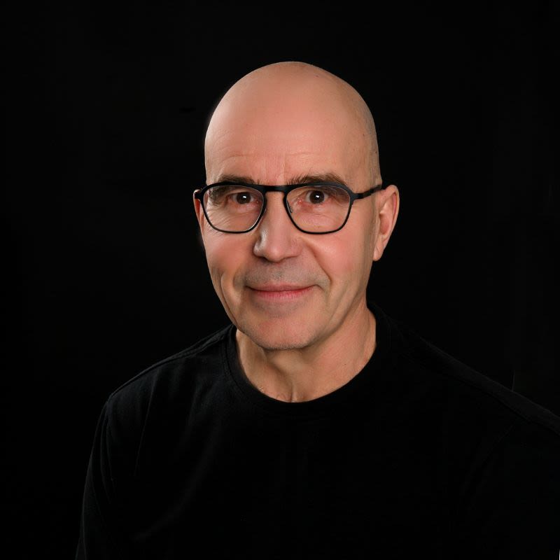 Mies ylävartalokuvassa kaljulla päällä ja mustalla t-paidalla tummassa studiotaustassa.