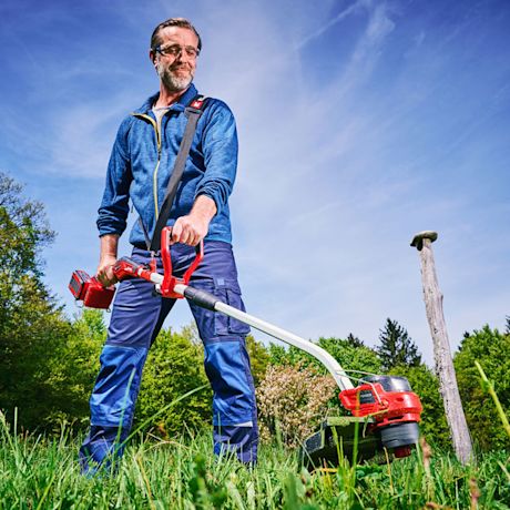 Mies sinisessä työpuvussa ajaa nurmikkoa punaisella akkutrimmerillä aurinkoisena päivänä.