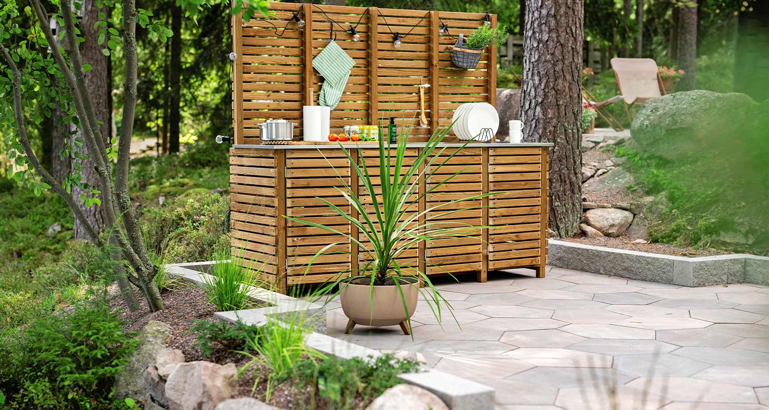 Puinen ulkokeittiö harmaalla laattaterassilla, ruukkuvehka ja vehreä metsänäkymä.