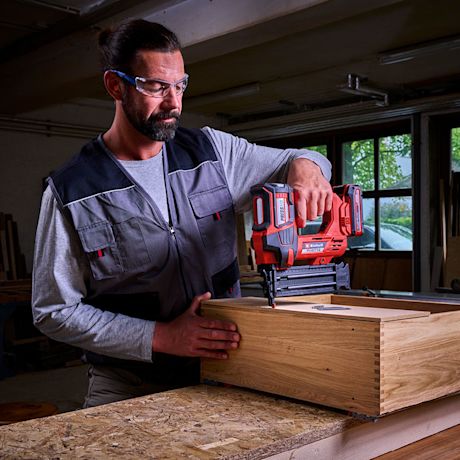 Työmies kiinnittää punaisella naulapyssyllä puisen laatikon liitoksia työpöydällä verstaassa.