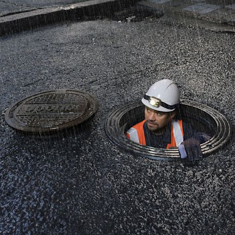 Sadekastelema asfalttitie, avoin viemärinluukku ja oranssiliivinen kypärätyömies sisällä