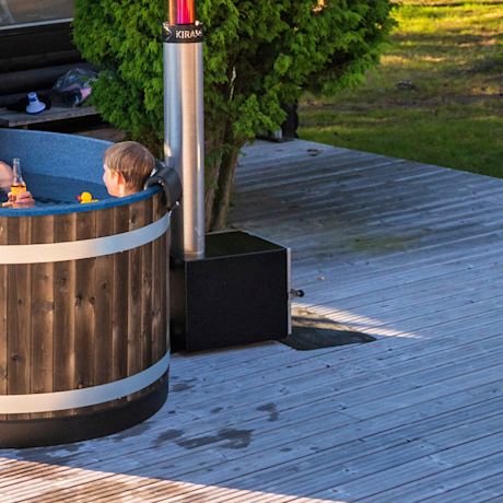 Lapsi sinisessä puukylpytynnyrissä, keltainen kumikylpyankka, terassi ja vihreät puut taustalla.