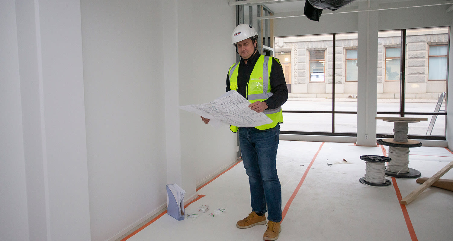 Rakennustyömaan sisätila, henkilö valkoisessa kypärässä ja keltaisessa liivissä pitelee piirustusta