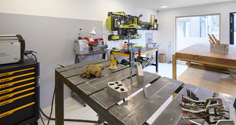 Kirkas metallityöpaja, keskipisteessä pöytiä, kelta-musta työkaluvaunu ja käsineet