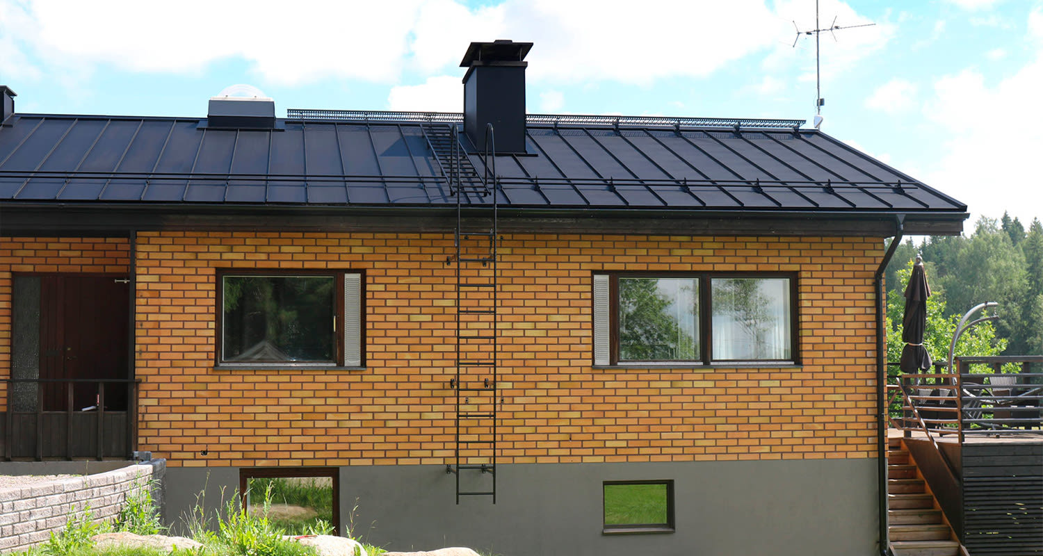 Oranssi tiilitalo mustalla peltikatolla tikkaineen, terassi kalusteineen ja metsä taustalla