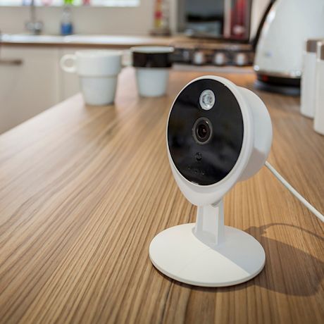 Valkoinen turvakamera mustalla etupaneelilla puupöydällä, keittiössä kahvikupit taustalla