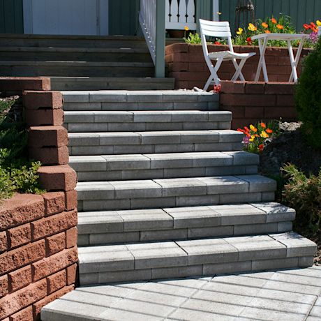 Harmaat betonportaat punatiiliseinämien välistä, ylhäällä valkoinen tuoli-pöytäryhmä ja kukkia.