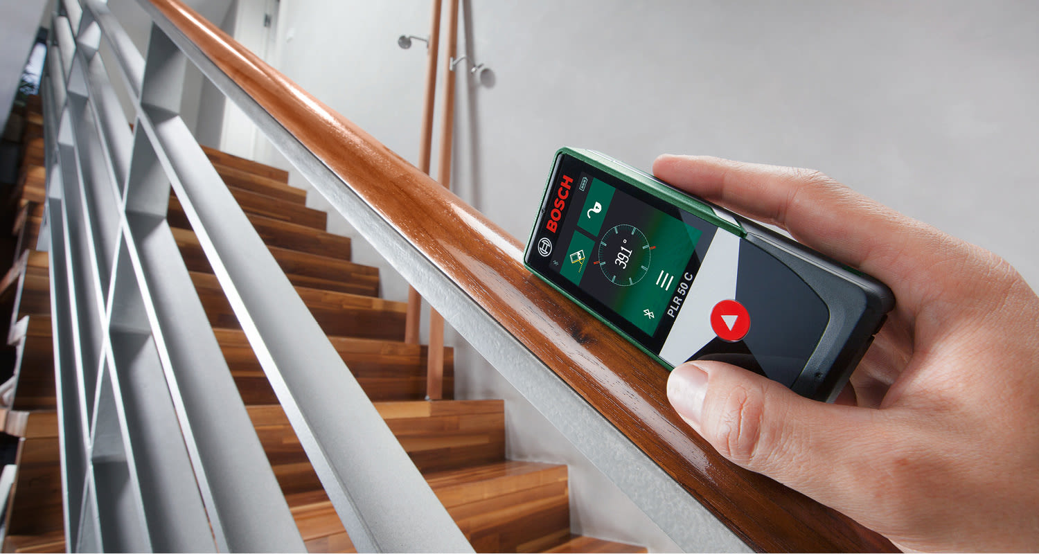 Käsi pitelee vihreä-harmaata Bosch PLR 50 C -laser-etäisyysmittaria puisen kaiteen yllä