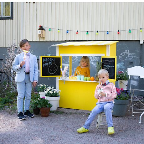 Pihalla keltainen juomakioski, lapset myyvät juomia ja hedelmiä, kukkia ja värivaloja.