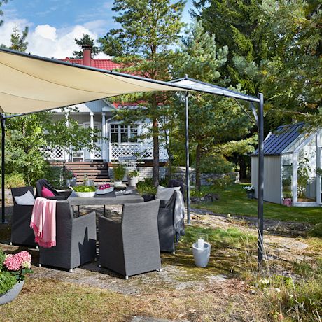 Harmaat puutarhatuolit ja pöytä beigen markiisin alla, pinkkejä tekstiilejä, valkoinen huvila