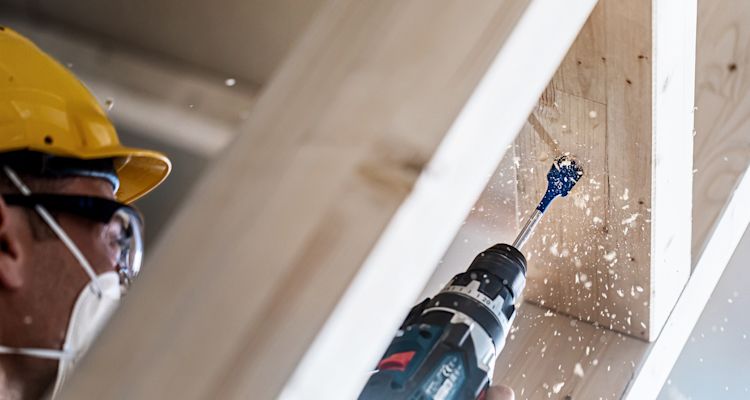 Rakennusmies keltakypärässä poraa alakaton puupalkkia sähköporalla, lastuja lentää.