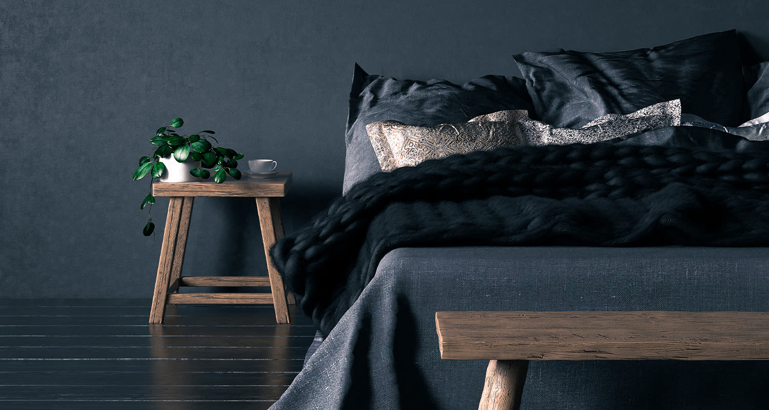 Tummanharmaa vuode mustalla viltillä, puupöytä viherkasvilla valkoisessa ruukussa ja kahvikupilla