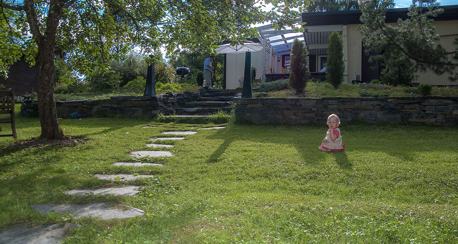 Vihreä piha kivipolkuineen, lapsi istuu nurmikolla, mies grillaa terassilla.