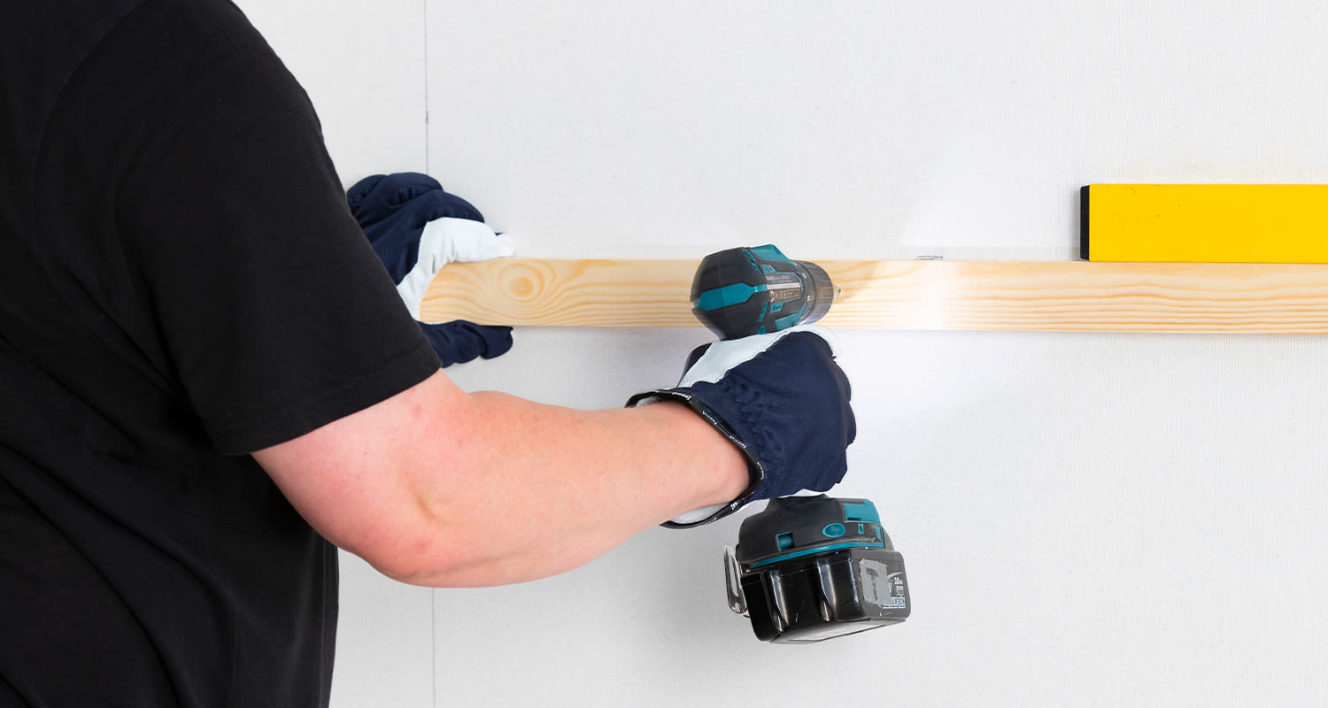 Käsineet kädessä porakoneella puulistaa seinään kiinnittävä henkilö keltaisen vatupassin vieressä.