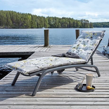 Harmaa kukkakuviollinen aurinkotuoli laiturilla järven äärellä, tarjottimella kannu, lasi ja päärynä.