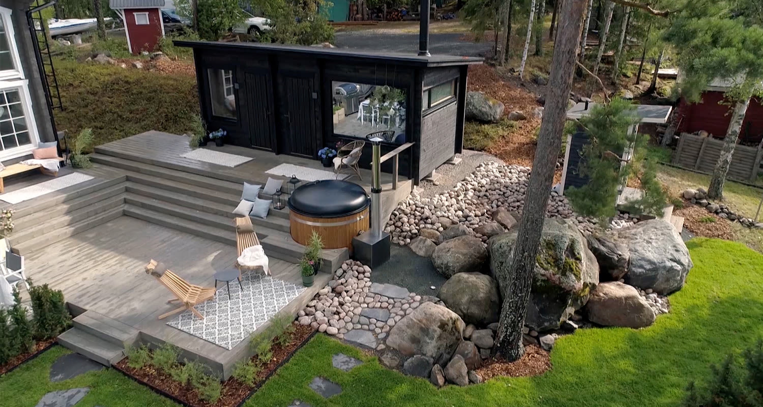 Harmaalankkuinen terassi puisilla tuoleilla, pyöreä kylpytynnyri ja musta saunamökki kalliopihoineen