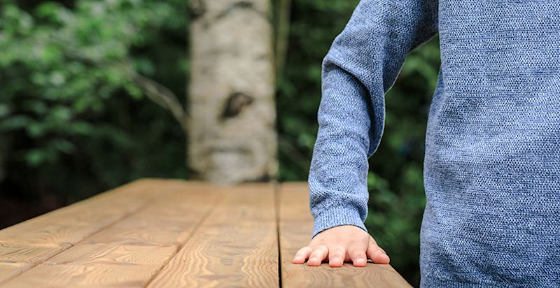 Sinineuleinen henkilö nojaa kädellä puiseen pöytään metsätaustan edessä.