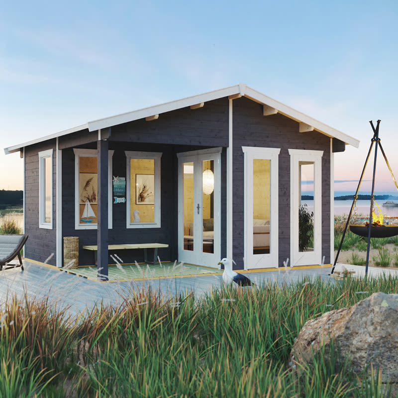 Musta hirsimökki valkoisin puittein rannalla, terassi, ruohoa ja lokki iltahämärässä.