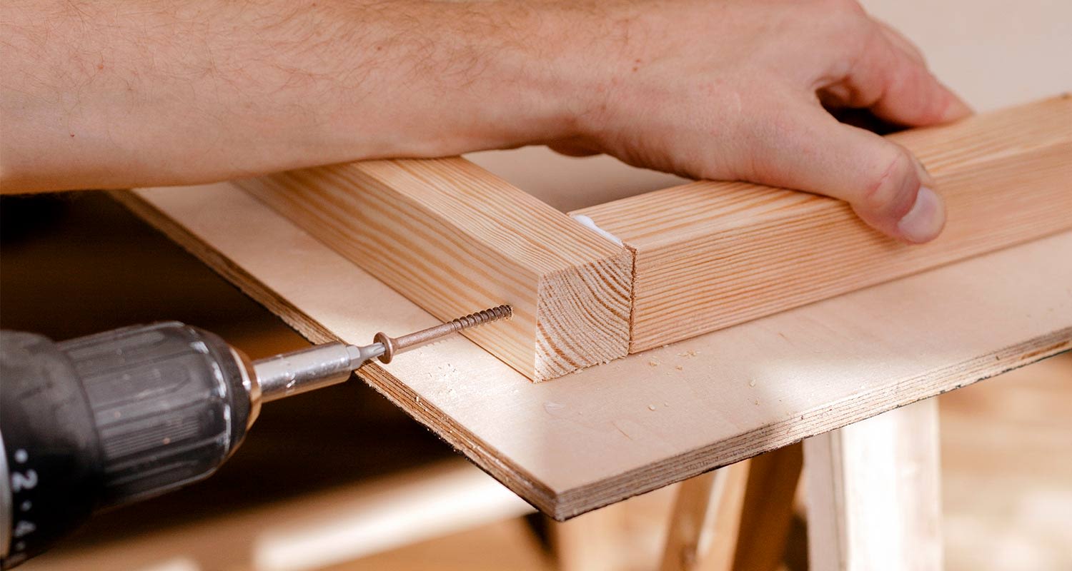 Käsi pitää vaaleaa puista kulmaliitosta vaneripöydällä, ruuvi porakoneen kärjessä.