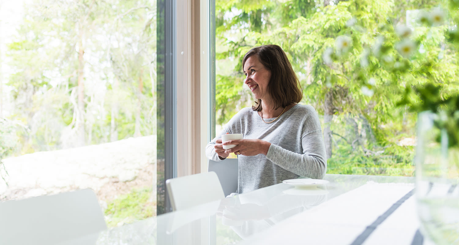 Henkilö harmaassa paidassa juo kahvia valkoisella pöydällä ja katsoo ikkunasta metsään.