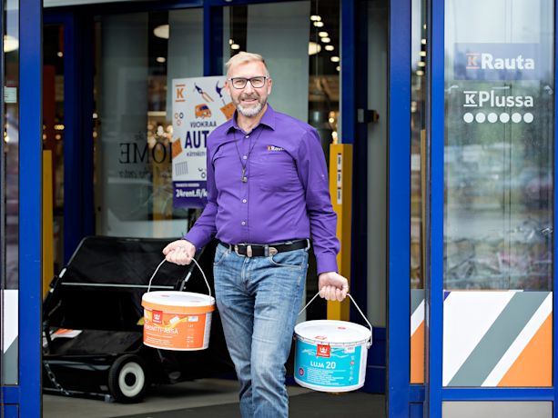 Sinisen oviaukon edessä violettipaitainen mies kantaa turkoosia ja oranssia maaliasäiliöitä