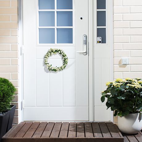 Valkoinen ulko-ovi sinisin ruuduin, vihreä kukkaseppele ja ruukkukasvit.