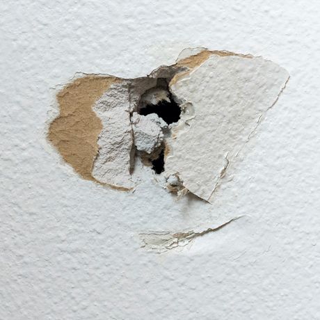 Valkoinen seinä, josta irronnut maali paljastaa ruskean kipsikerroksen ja mustan reiän.