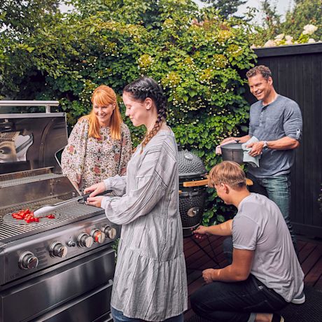 Neljä ihmistä puutarhassa grillaamassa tomaatteja kaasugrillissä, taustalla vihreää pensasta.