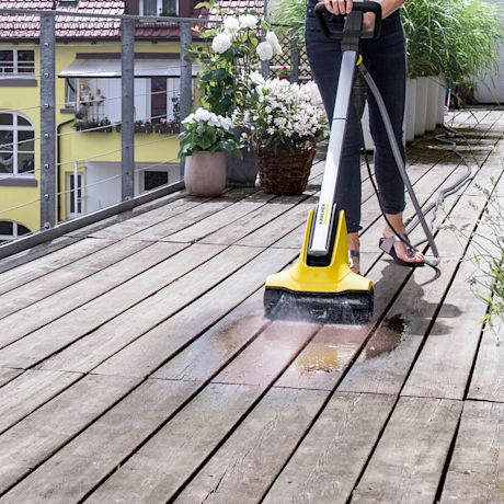 Henkilö pesee keltaisella Kärcher-puhdistuskoneella puisella parvekkeella kukkaruukkujen vieressä.