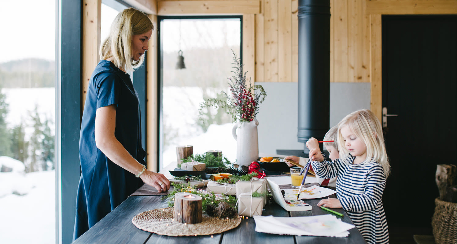 Puupöydällä paketit, vihreät havut ja punaiset marjat, lapset maalaavat ja aikuinen ikkunan ääressä.