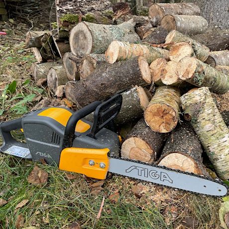 Stiga CS 700E -akkuketjusaha puupinon edessä ulkona, koivupuita ja muuta puuta sahattuna.