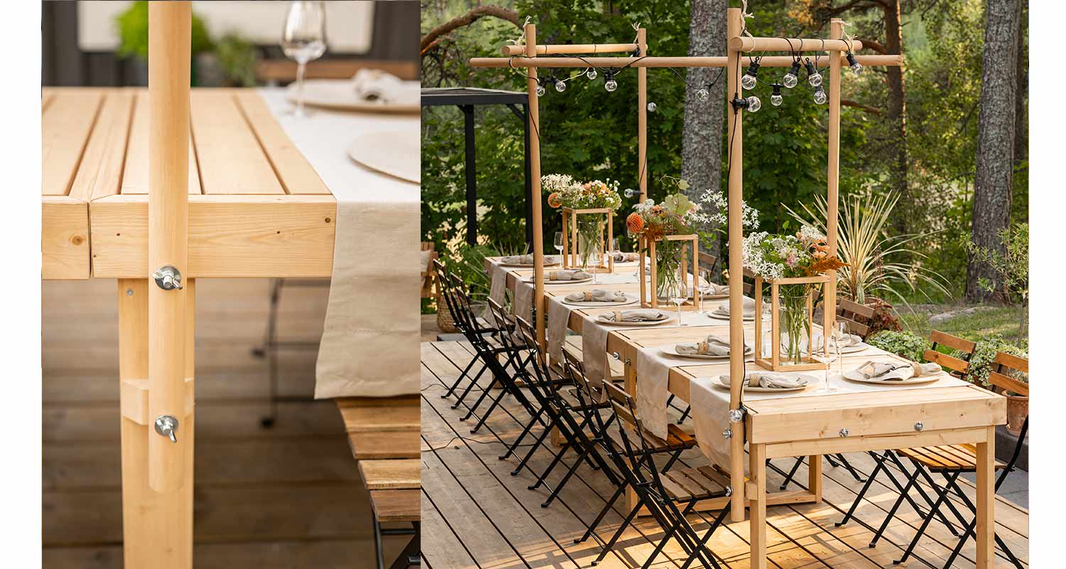 Ulkoilmajuhlapöytä vaaleasta puusta, beige liina, kukat, mustat tuolit ja valosarja puutarhassa