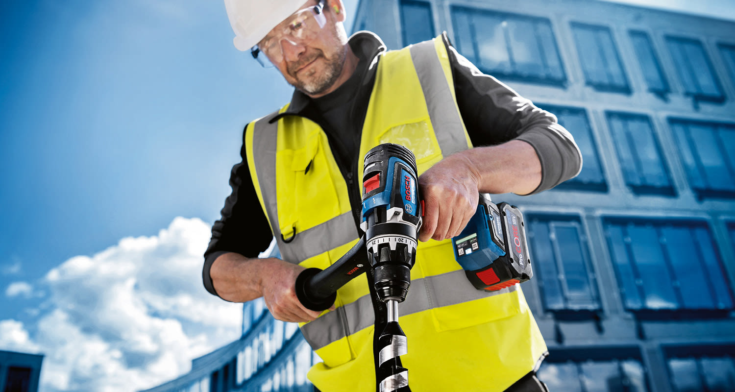 Työmies keltaisessa suojaliivissä ja kypärässä käyttää Bosch-porakonetta lasirakennuksen edessä