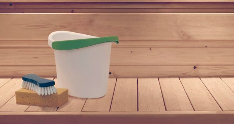 Valkoinen ämpäri vihreällä reunuksella, sininen harja keltaisen sienen päällä puupinnalla.