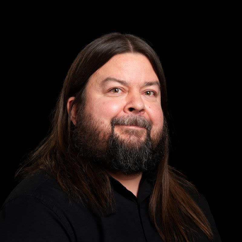 Pitkähiuksinen mies kastanjanruskeilla hiuksilla ja täyteläisellä parralla mustaa taustaa vasten.