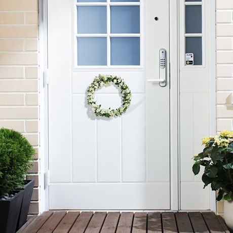 Valkoinen ulko-ovi sinisillä yläikkunoilla, kukkaseppele ja ruukkukasvit.