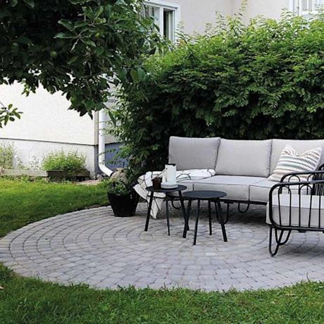 Harmaa kulmasohva ja mustat sivupöydät pyöreällä harmaalla laattapatiolla taustalla vihreää pensasta