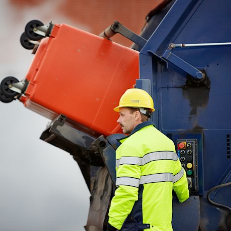 Jäteauto nostaa punaista roskista, työntekijä keltaisessa kypärässä ja heijastinliivissä