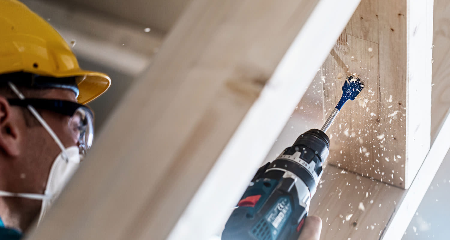 Keltakypäräinen poraa sinisellä terällä vaaleaa puupalkkia, puujauhoa lentää.