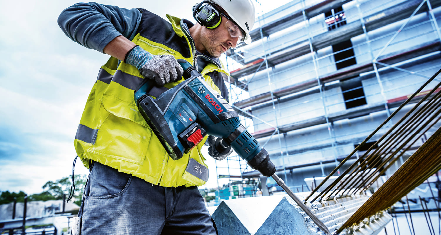 Rakennustyömies liivissä poraa sinisellä vasaraporakoneella rakennustelineiden edessä