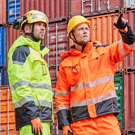Työmiehet keltaisissa ja oransseissa suojavaatteissa konttien edessä, toinen osoittaa.