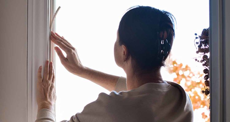 Ruskeahiuksinen nainen avaa valoa tulvivaa ikkunaa, taustalla oransseja syyslehtiä.