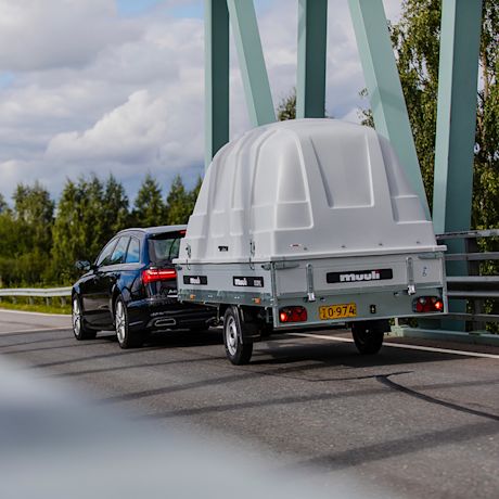 Musta farmariauto vetää valkoharmaata kupukattoista peräkärryä vihreällä sillalla, taustalla metsä