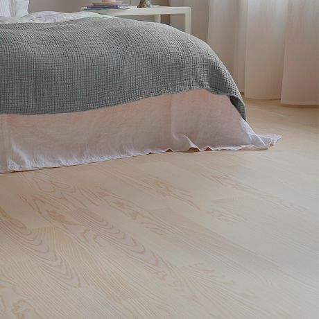 Valoisassa makuuhuoneessa vaalea puulattia, valkoinen lakanakangas ja harmaa täkki.